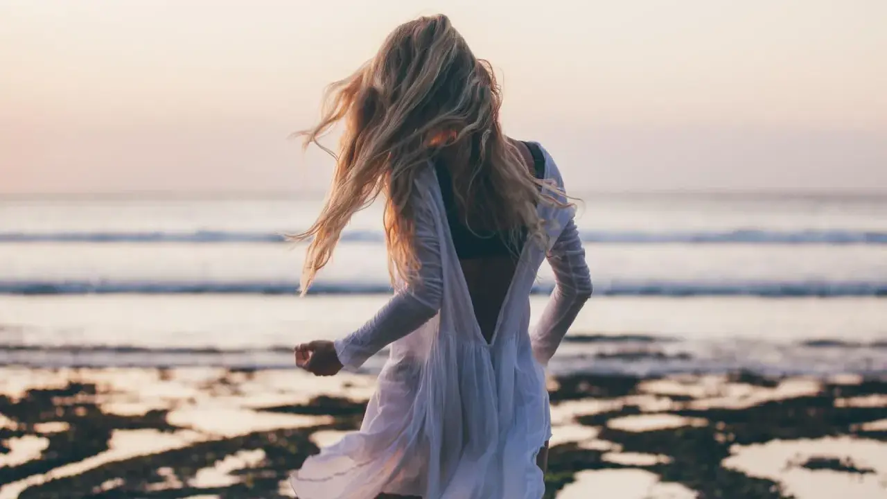 Blondynka w białej sukience tańczy na plaży, wiatr rozwiewa jej włosy, a w tle widać spokojne beach waves.