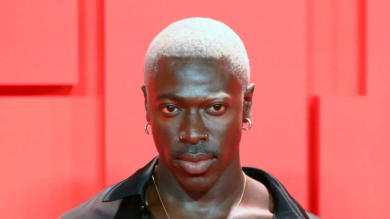Mężczyzna z buzz cut blond, kolczykami w uszach i nosie, na tle czerwonych elementów.