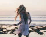 Blondynka w białej sukience tańczy na plaży, wiatr rozwiewa jej włosy, a w tle widać spokojne beach waves.