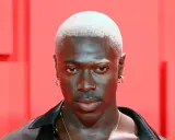 Mężczyzna z buzz cut blond, kolczykami w uszach i nosie, na tle czerwonych elementów.