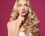 Zastanawiasz się, jak sprawdzić czy pasuje Ci blond? Kobieta z burzą złotych loków na różowym tle z palcem przy ustach.