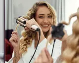Uśmiechnięta blondynka tworzy fale na długich włosach za pomocą lokówki.