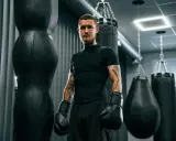 Mężczyzna w rękawicach bokserskich, z tatuażami, stoi obok worków treningowych. Jego krótka fryzura, niczym fryzura na zapałkę damska, podkreśla jego determinację.