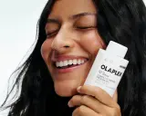 Uśmiechnięta kobieta trzyma butelkę Olaplex N°3 Plus. Dowiedz się, jak stosować Olaplex 0 dla idealnych włosów.