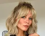 Kobieta z niebieskimi oczami i piegami, z falowanymi blond włosami i cieniowaną grzywką, upiętymi w luźny kok.