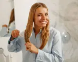 Uśmiechnięta blondynka wykonuje pielęgnację włosów krok po kroku, szczotkując swoje długie, jasne pasma.