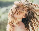Kobieta z burzą blond loków, które opadają na twarz. Jej uśmiech i oczy są częściowo ukryte, ale widać, że cieszy się swobodą. To efekt głębokiego nawilżenia włosów kręconych.