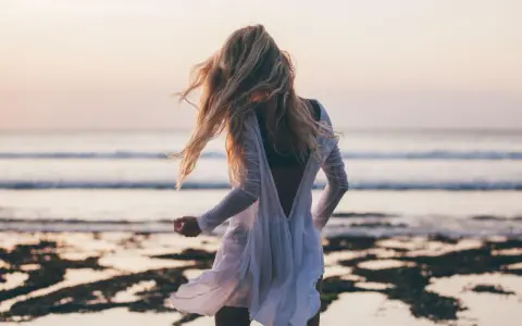 Blondynka w białej sukience tańczy na plaży, wiatr rozwiewa jej włosy, a w tle widać spokojne beach waves.