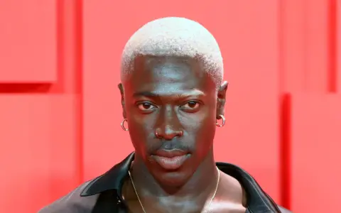 Mężczyzna z buzz cut blond, kolczykami w uszach i nosie, na tle czerwonych elementów.