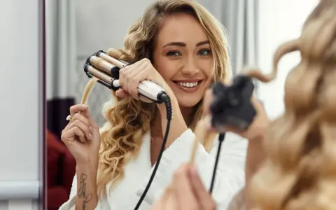 Uśmiechnięta blondynka tworzy fale na długich włosach za pomocą lokówki.