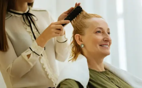 Fryzjer do kręconych włosów czesze blondynkę. Kobieta uśmiecha się, czując się komfortowo.