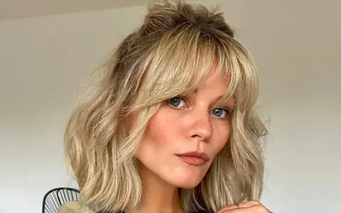 Kobieta z niebieskimi oczami i piegami, z falowanymi blond włosami i cieniowaną grzywką, upiętymi w luźny kok.