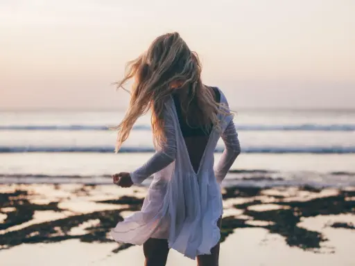 Blondynka w białej sukience tańczy na plaży, wiatr rozwiewa jej włosy, a w tle widać spokojne beach waves.