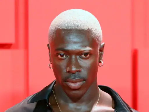 Mężczyzna z buzz cut blond, kolczykami w uszach i nosie, na tle czerwonych elementów.