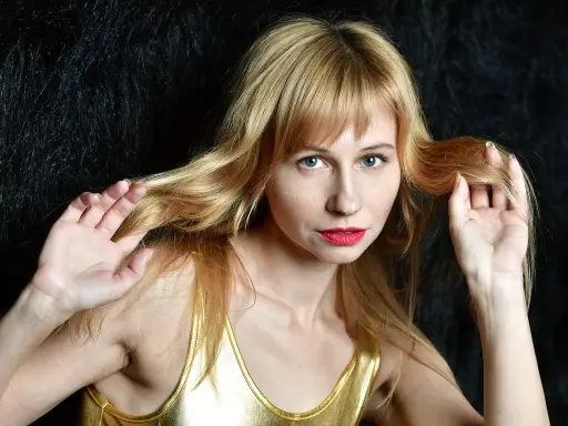 Blondynka w złotym topie, z grzywką, która odrasta, bawi się włosami na tle czarnego futra.