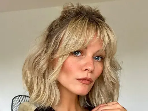 Kobieta z niebieskimi oczami i piegami, z falowanymi blond włosami i cieniowaną grzywką, upiętymi w luźny kok.