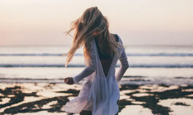 Blondynka w białej sukience tańczy na plaży, wiatr rozwiewa jej włosy, a w tle widać spokojne beach waves.