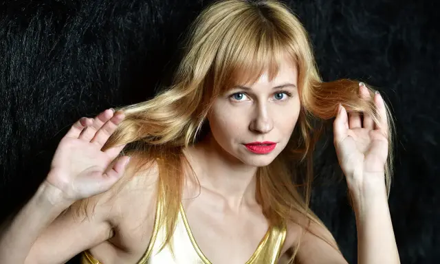 Blondynka w złotym topie, z grzywką, która odrasta, bawi się włosami na tle czarnego futra.