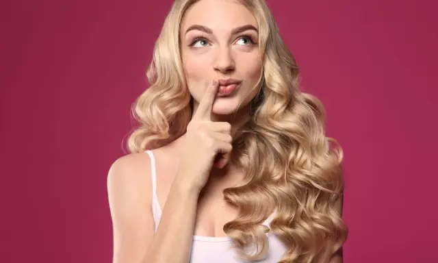 Zastanawiasz się, jak sprawdzić czy pasuje Ci blond? Kobieta z burzą złotych loków na różowym tle z palcem przy ustach.