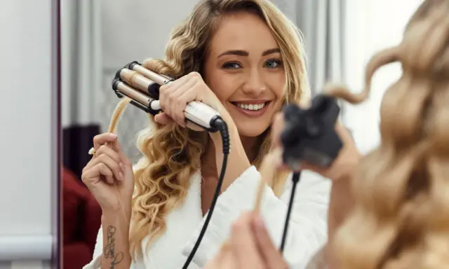Uśmiechnięta blondynka tworzy fale na długich włosach za pomocą lokówki.