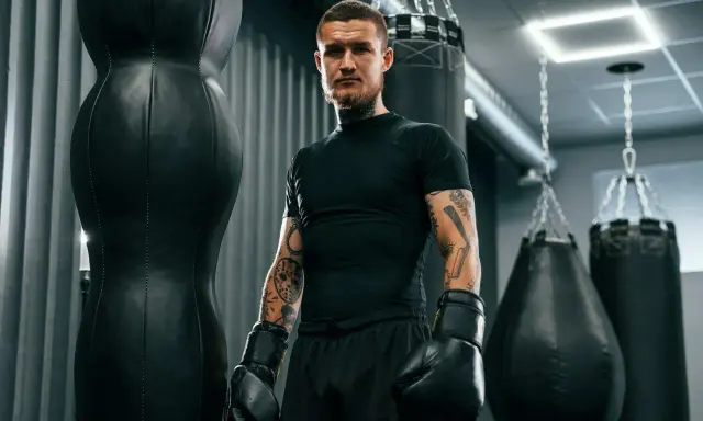 Mężczyzna w rękawicach bokserskich, z tatuażami, stoi obok worków treningowych. Jego krótka fryzura, niczym fryzura na zapałkę damska, podkreśla jego determinację.