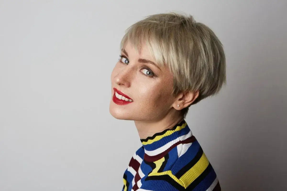 Kobieta z krótkimi blond włosami w stylu pixie bob, z rudymi ustami i w kolorowym swetrze. Ta fryzura pixie bob dla kogo? Dla odważnych!